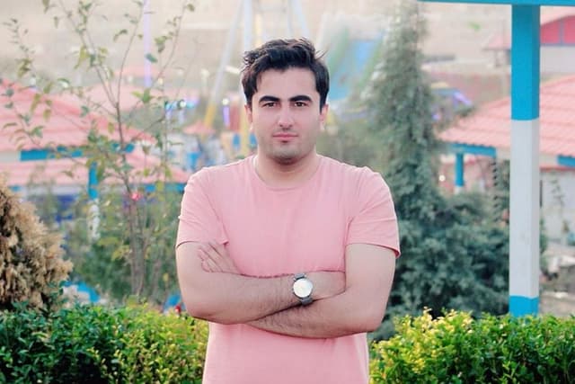 Headshot photo of a young man standing with his arms crossed, with trees and buildings in the background.