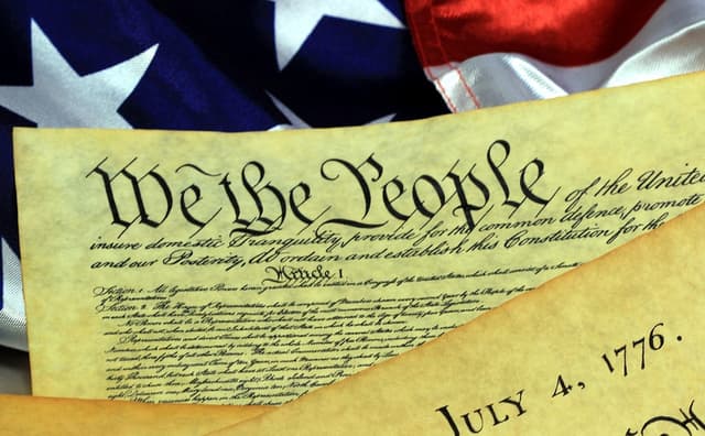 A snippet of the U.S. constitution with the words, “We the People,” and the American flag in the background.