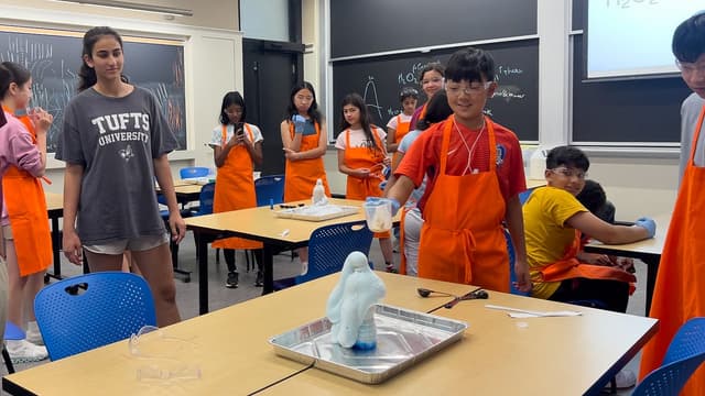 Candid photo of a classroom with ten middle school students wearing aprons and safety goggles looking at a chemistry experiment on the table, where foam is bubbling out of a water bottle.