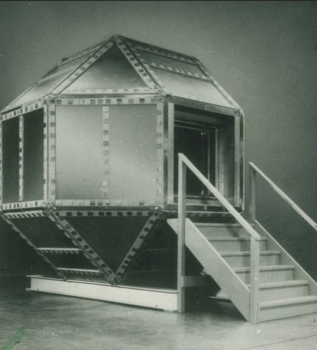 A black and white photo of a shielded hexagonal metal chamber atop metal girders.